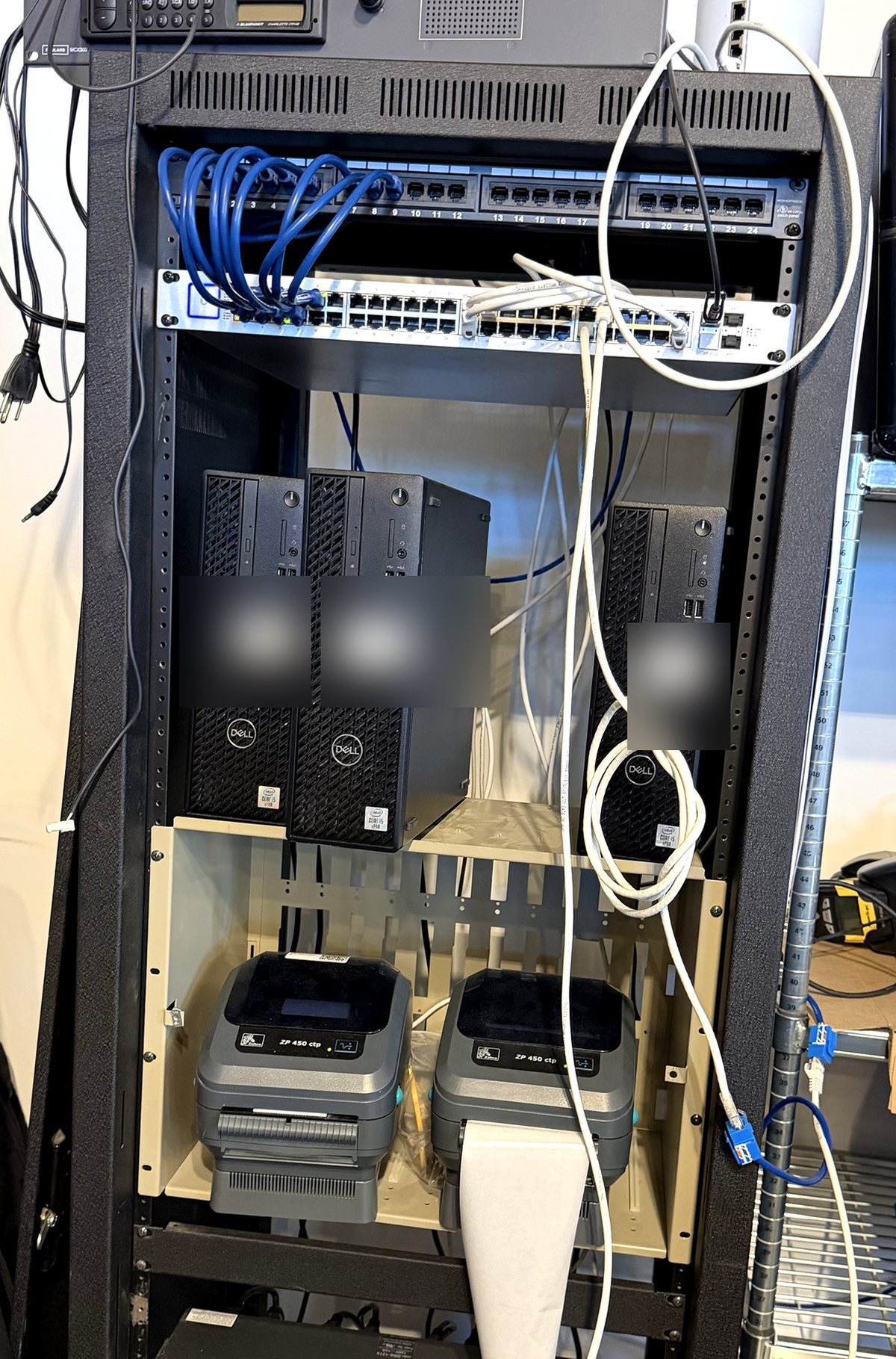 The office rack: patch panel and switch up top, three small-form-factor Dell OptiPlex Proxmox hosts in the middle with their labels blurred, and two thermal label printers on the shelf below