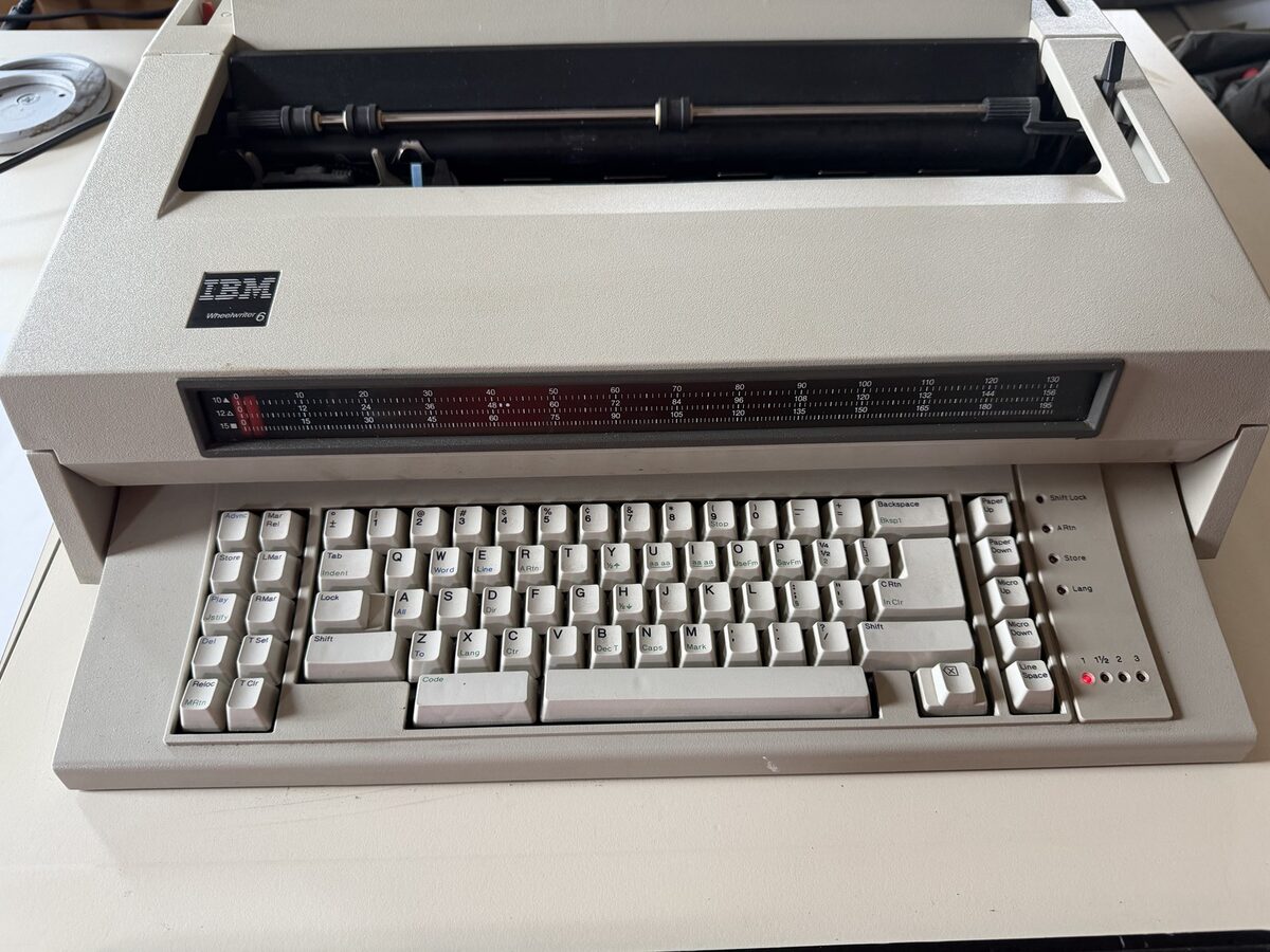 A beige IBM Wheelwriter 6 electronic typewriter with its top cover lifted, exposing the platen roller and carriage rail above the keyboard