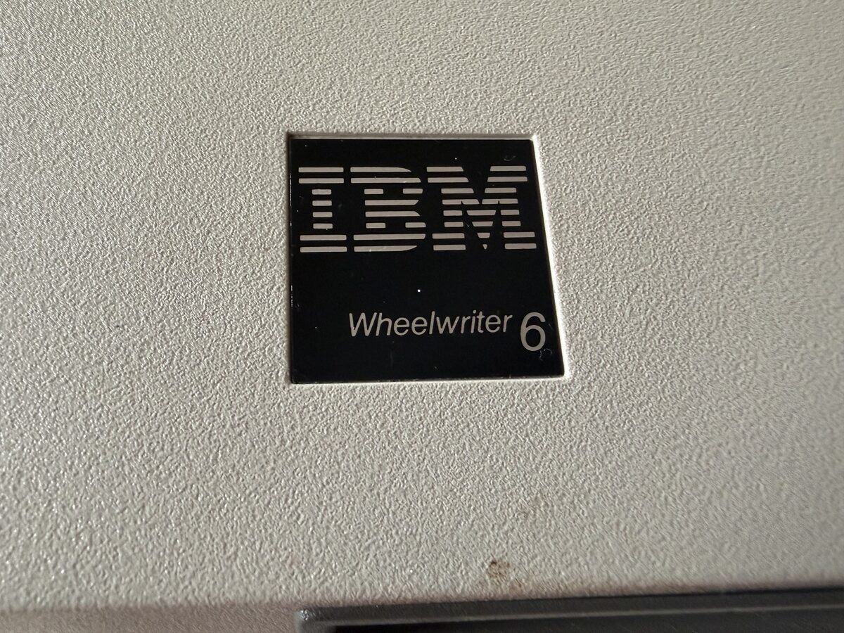 Close-up of a black IBM Wheelwriter 6 badge on a beige plastic machine housing, the model number legible against the textured panel